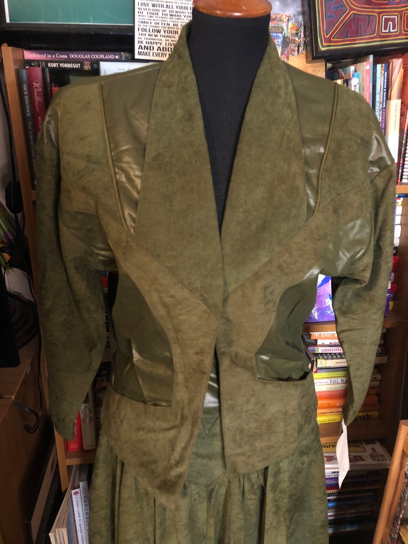 May include: A green suede jacket and skirt set with a faux leather trim. The jacket has a V-neck and long sleeves. The skirt is a-line and has a pleated design.