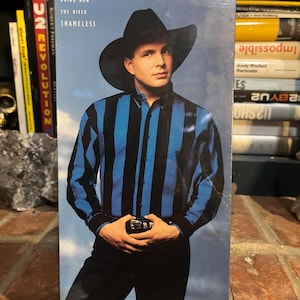 May include: A Garth Brooks Ropin' The Wind VHS tape featuring a cover image of a person wearing a black cowboy hat and a blue and black vertical striped long-sleeve shirt. The text includes song titles like Rodeo, Papa Loved Mama, and The River.