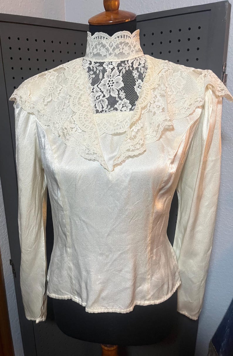 May include: Ivory-colored blouse with long sleeves and a high, lace-trimmed collar. The blouse features a satin-like fabric and a delicate lace insert at the neckline. The vintage-style top is displayed on a mannequin.