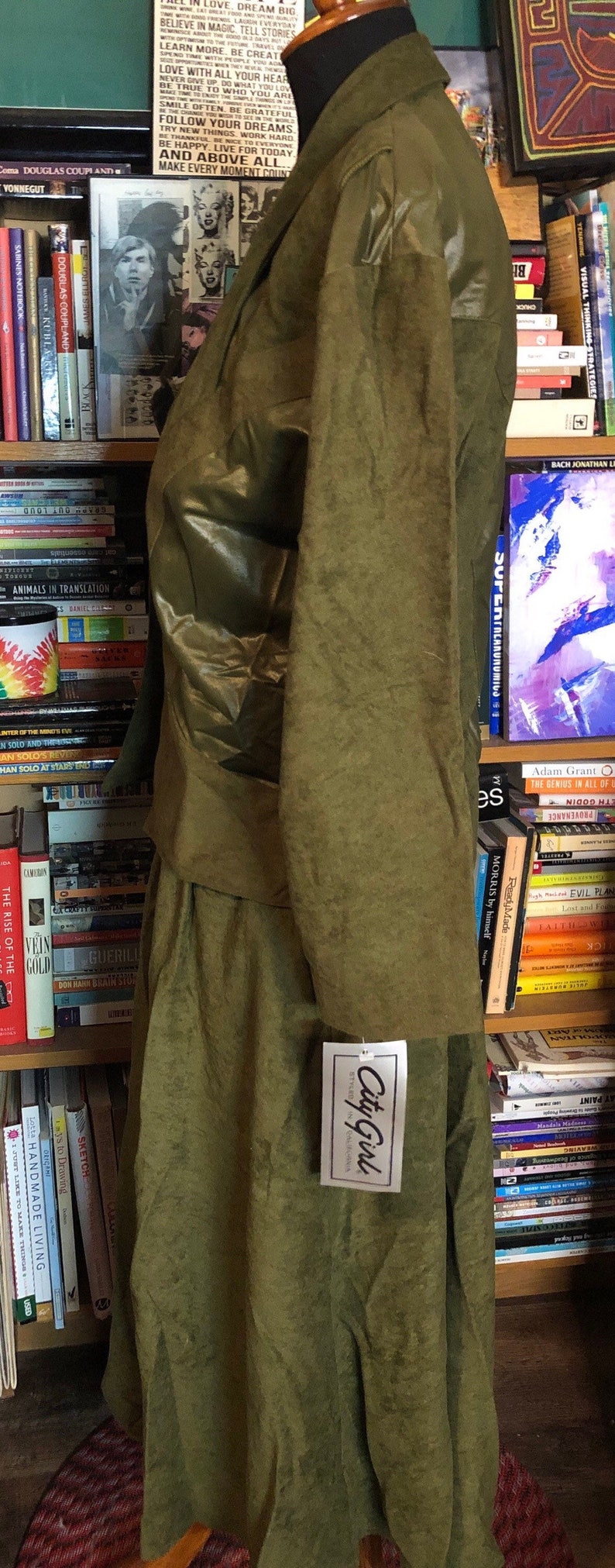 May include: A green, long-sleeved jacket with a matching skirt. The jacket has a shiny, almost metallic, finish. The skirt is a more matte fabric. The outfit is displayed on a mannequin in front of a bookshelf. A tag with the text "City Girl" is attached to the skirt.