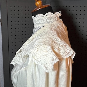 May include: Ivory-colored vintage dress with a high, lace-trimmed collar. The dress is displayed on a mannequin, showcasing the delicate lace detailing and the soft, flowing fabric. The dress appears to be made of a lightweight material.