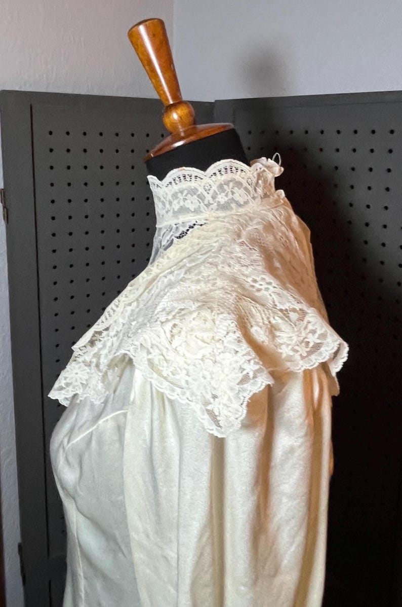 May include: Ivory-colored vintage dress with a high, lace-trimmed collar. The dress is displayed on a mannequin, showcasing the delicate lace detailing and the soft, flowing fabric. The dress appears to be made of a lightweight material.
