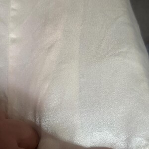 May include: Close-up of a cream-colored satin fabric. The fabric has a subtle sheen and a smooth texture, with visible folds and creases. The image highlights the fabric's delicate appearance and the way it catches the light.