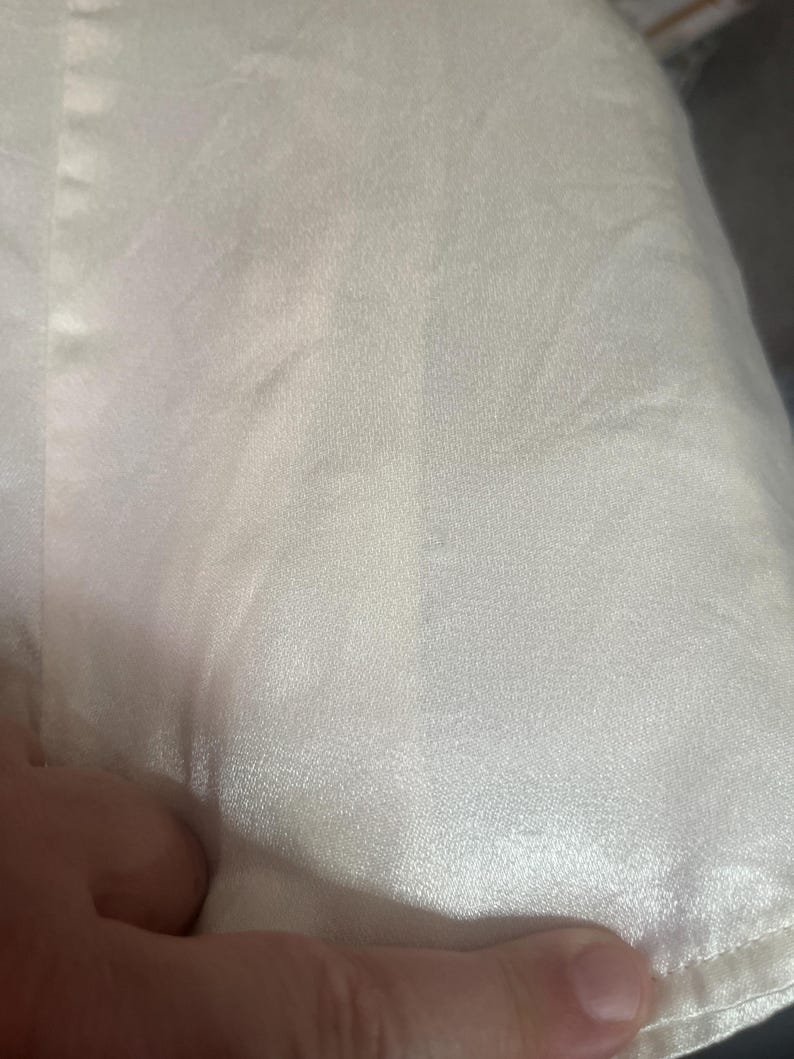 May include: Close-up of a cream-colored satin fabric. The fabric has a subtle sheen and a smooth texture, with visible folds and creases. The image highlights the fabric's delicate appearance and the way it catches the light.