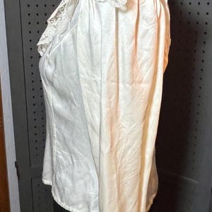May include: Ivory-colored blouse with a high, ruffled lace collar. The long-sleeved top is made of a silky fabric and is displayed on a mannequin. The blouse has a vintage or antique aesthetic.