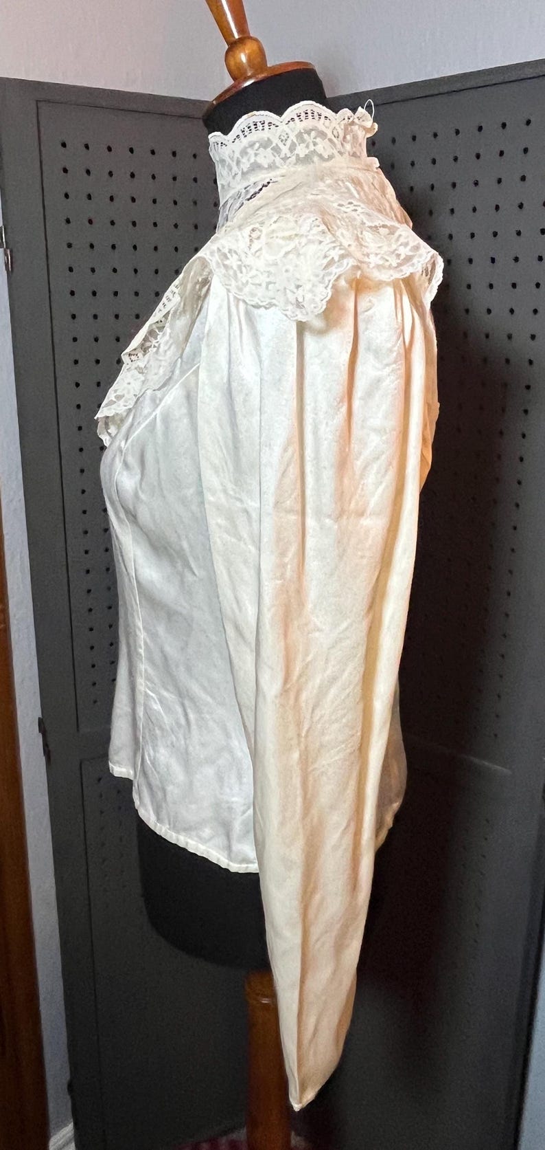 May include: Ivory-colored blouse with a high, ruffled lace collar. The long-sleeved top is made of a silky fabric and is displayed on a mannequin. The blouse has a vintage or antique aesthetic.