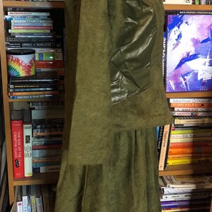 May include: A long, green, suede jacket with a large, shiny, green patch on the left sleeve. The jacket has a collar and a long skirt.