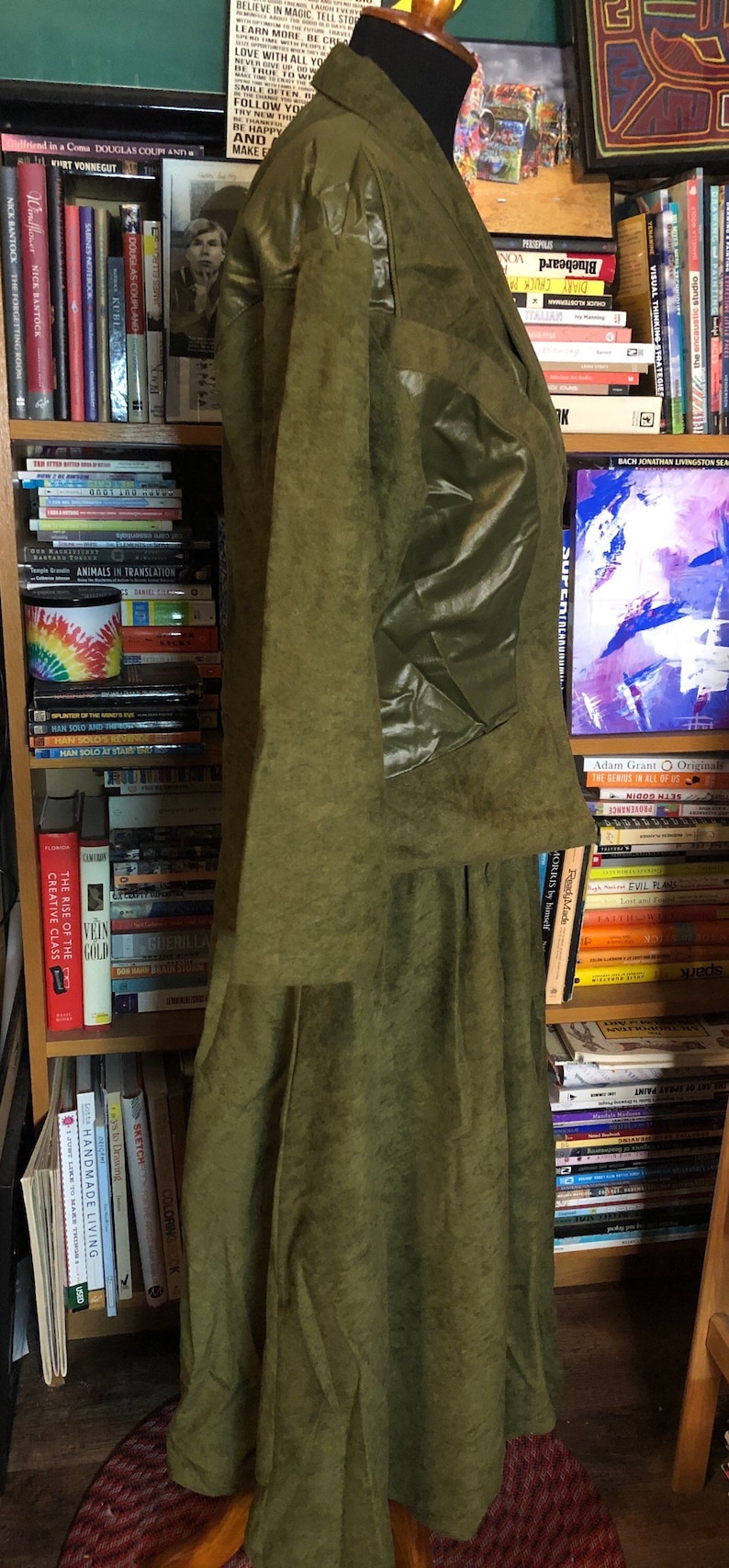 May include: A long, green, suede jacket with a large, shiny, green patch on the left sleeve. The jacket has a collar and a long skirt.