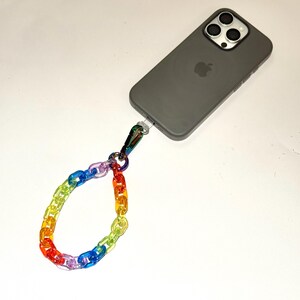 Puede incluir: Un smartphone gris con una correa de muñeca de cadena de arcoíris. Los eslabones de la cadena transitan por los colores del arcoíris, sujetos al teléfono con un cierre plateado. El teléfono tiene una cámara de triple lente.