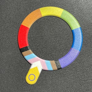 Könnte beinhalten: Ein bunter, kreisförmiger, regenbogenfarbener Schlüsselanhänger mit einem gelben Griff und einem kleinen Kreis in der Mitte. Der Schlüsselanhänger besteht aus mehreren Farben, darunter Rot, Orange, Gelb, Grün, Blau, Lila und Schwarz.