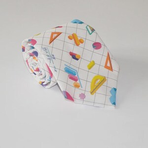 May include: White necktie with a grid pattern and colorful geometric shapes, including triangles, pie charts, and rulers. The tie is rolled up, showcasing the patterned fabric.