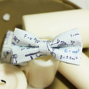 Puede incluir: Una pajarita blanca con un patrón de cuadrícula azul y fórmulas científicas escritas a mano, incluyendo "F=ma", "E=mc²" y "g=9.8ms²".
