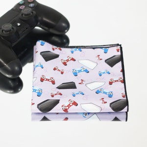 Könnte beinhalten: Ein lila Mikrofaser-Reinigungstuch mit einem sich wiederholenden Muster aus Videospielcontrollern, Spielkonsolen und X und O. Das Tuch ist in der Hälfte gefaltet und liegt auf einer weißen Oberfläche.