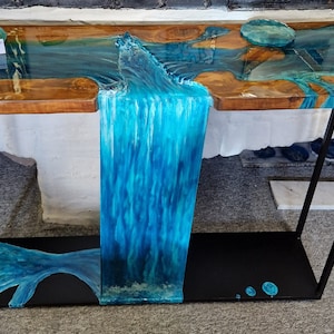 May include: A wooden table with a black metal frame. The tabletop is made of wood and epoxy resin, with a blue and white waterfall design. The table has a shelf underneath.