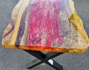 May include: A wooden table with a live edge top and a black metal X-shaped base. The top is finished with a clear epoxy resin that has a pink and orange swirl pattern.