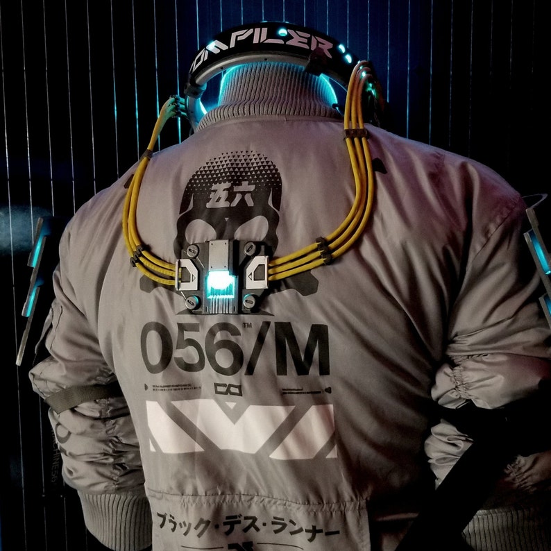 Puede incluir: Una chaqueta gris con una correa amarilla y negra cruzada sobre el pecho. La correa tiene una luz azul y el texto "056/M" est&aacute; impreso en la chaqueta. La chaqueta tiene un gr&aacute;fico de calavera con el texto japon&eacute;s "左" en la parte delantera.