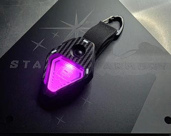 Puede incluir: Un llavero negro y de fibra de carbono con una luz rosa brillante. El llavero está sujeto a una correa negra y a un mosquetón plateado. El llavero está sobre una superficie triangular negra con el texto "STAR ARMORY".