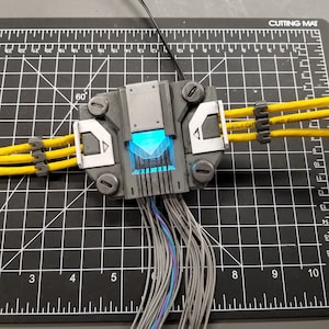 Puede incluir: Una caja gris y blanca impresa en 3D con una luz azul en su interior. La caja tiene varios cables que salen de ella, algunos de los cuales est&aacute;n conectados a cables amarillos con bridas de cable negras. La caja est&aacute; sentada en una estera de corte negra con marcas blancas.