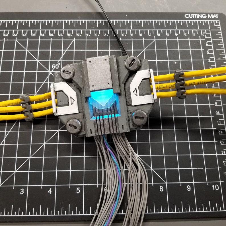 Puede incluir: Una caja gris y blanca impresa en 3D con una luz azul en su interior. La caja tiene varios cables que salen de ella, algunos de los cuales est&aacute;n conectados a cables amarillos con bridas de cable negras. La caja est&aacute; sentada en una estera de corte negra con marcas blancas.