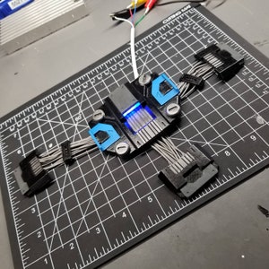 Puede incluir: Un dispositivo impreso en 3D negro y azul con varios cables grises conectados a &eacute;l. El dispositivo tiene una luz azul que brilla a trav&eacute;s de &eacute;l.