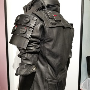 May include: Black leather jacket with a zipper closure and a black, padded, and textured shoulder piece. The jacket has a black, rectangular, textured patch on the back with the text "TIME - 1.1.1A".