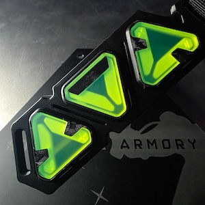 Puede incluir: Dispositivo rectangular negro con tres secciones triangulares translúcidas de color verde neón. El dispositivo tiene un acabado mate y está montado sobre una superficie negra. La palabra "ARMORY" es visible.