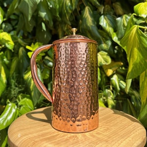 May include: A copper-colored pitcher with a hammered texture, a curved handle, and a lid with a small knob. The pitcher is cylindrical and sits on a wooden surface, with green foliage in the background.