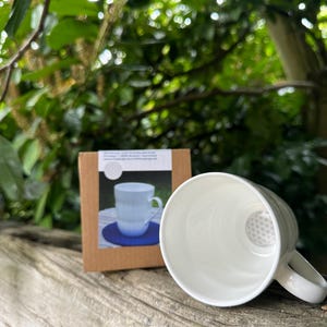 May include: A white ceramic tea cup with a built-in infuser, resting on a wooden surface. A cardboard box with a picture of the cup is next to it. The background features lush green foliage.