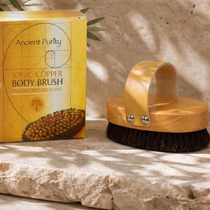 Puede incluir: Una caja rectangular amarilla etiquetada como Ancient Purity Ionic Copper Body Brush junto a un cepillo corporal de madera con cerdas oscuras y una correa de cuero beige asegurada por dos remaches de metal plateado.