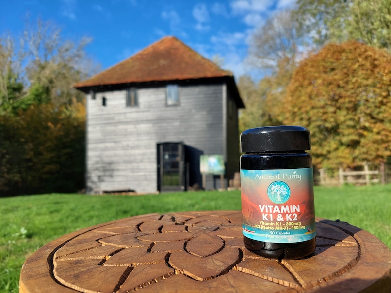 May include: A black plastic bottle of Ancient Purity Vitamin K1 & K2 dietary supplement capsules. The label features a gradient background, a tree logo, and text stating Vitamin K1 - 300mcg and K2 (Natto MK-7) - 150mcg with 90 capsules total.