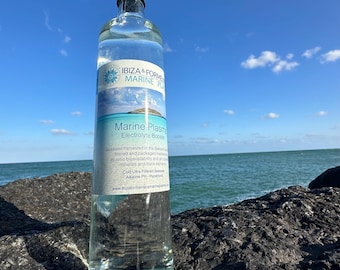 Puede incluir: Una botella de vidrio transparente de Marine Plasma Electrolyte Booster, un suplemento a base de agua de mar. La botella está sentada sobre una superficie rocosa con un océano azul y cielo de fondo. La etiqueta presenta el nombre de la marca "IBIZA & FORMENTERA MARINE PLASMA" y el nombre del producto "Marine Plasma Electrolyte Booster".