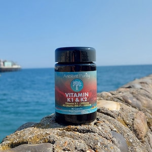 May include: A black plastic bottle of Ancient Purity Vitamin K1 & K2 food supplement with 90 capsules. The label features a tree logo, red and blue gradient background, and text detailing Vitamin K1 300mcg and K2 (Natto MK-7) 150mcg content.