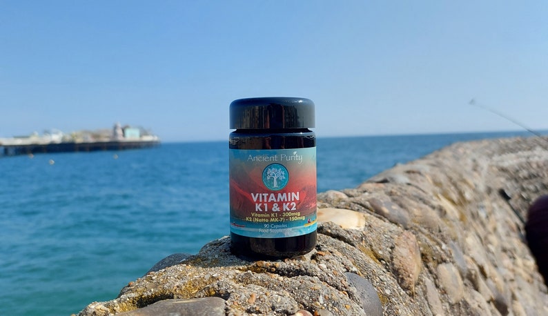 May include: A black plastic bottle of Ancient Purity Vitamin K1 & K2 food supplement with 90 capsules. The label features a tree logo, red and blue gradient background, and text detailing Vitamin K1 300mcg and K2 (Natto MK-7) 150mcg content.