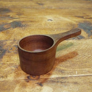 Peut inclure: Une louche en bois avec une poignée, fabriquée en bois brun foncé. La louche a une forme de coupe cylindrique et une poignée qui s'étend sur le côté. Elle est probablement utilisée pour mesurer des ingrédients secs.