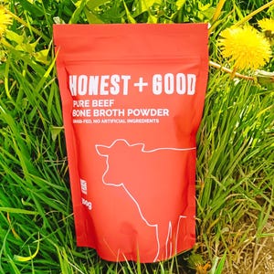 Peut inclure: Pochette rouge avec du texte blanc qui dit "HONEST+GOOD PURE BEEF BONE BROTH POWDER GRASS-FED, NO ARTIFICIAL INGREDIENTS" et un dessin au trait d'une vache. La pochette est posée sur un lit d'herbe verte.