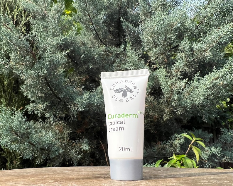 May include: A white tube of Curaderm topical cream with a gray cap, against a backdrop of green foliage. The tube displays the brand name and product details in green and black text, including the 20ml volume.