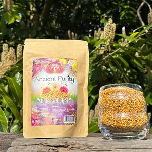 May include: A brown paper bag with the text "Ancient Purity" and "Bee Pollen" printed on it. The bag is sitting on a wooden surface next to a glass jar filled with yellow bee pollen.