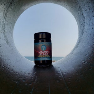 May include: A dark-colored supplement bottle with a black cap and a label featuring a tree logo, the text Vitamin K1 & K2, and additional supplement details. The bottle is positioned within a circular opening that frames the product against a sky.