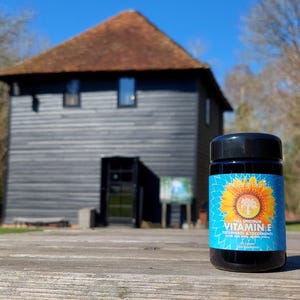 May include: A blue and yellow jar of "Nature's Truth" full spectrum Vitamin E supplement with tocopherols and tocotrienols. The jar is sitting on a wooden surface in front of a wooden building.