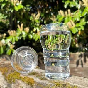 May include: Two clear glass shot glasses, one with a textured exterior and the other with a smooth exterior, both filled with water. The glasses are sitting on a wooden surface with moss growing on it.