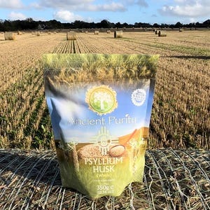 Puede incluir: Una bolsa de Ancient Purity Psyllium Husk en un campo de pacas de heno. La bolsa es blanca con detalles verdes y presenta un logotipo de árbol. El texto en la bolsa dice "Ancient Purity Psyllium Husk 350g (12.3 oz)".