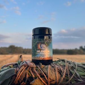 May include: A black supplement bottle with a label that reads "Selenium" and "90 Tablets". The label features a tree graphic and the words "Ancient Purity". The bottle sits on a bundle of dried herbs, with a field and sky in the background.