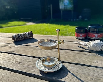 May include: A brass incense burner with a round base and a mesh top. The burner is on a wooden table with two bundles of sage and two jars of "Ancient Purity" incense.