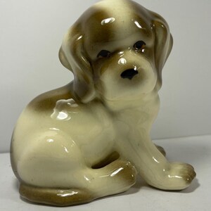May include: A ceramic puppy figurine in a sitting position. The puppy has a cream-colored body with brown markings on its head, ears, and paws. It has dark eyes and a black nose. The figurine is glossy and appears to be vintage.