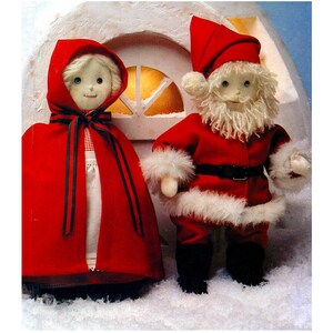 May include: A red-hooded doll and a Santa Claus doll stand in front of a white house. The Santa doll has a white beard and a black belt. The dolls are made of fabric and have a vintage look.