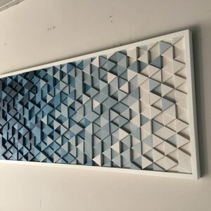 Peut inclure: Une &oelig;uvre d'art murale en bois avec un d&eacute;grad&eacute; de triangles bleus. Les triangles sont dispos&eacute;s en un motif g&eacute;om&eacute;trique et sont peints dans des nuances de bleu, du bleu fonc&eacute; au bleu clair. La pi&egrave;ce est encadr&eacute;e de blanc.