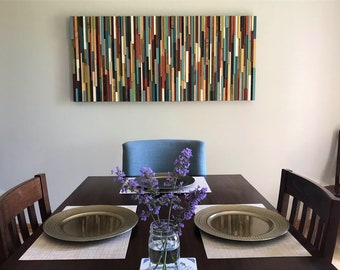 May include: A wooden wall art piece with vertical strips of wood in various colors, including blue, green, red, yellow, and brown. The art is hanging on a white wall above a dining table with four chairs.