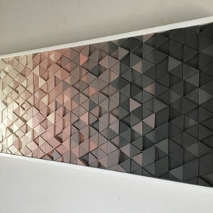 May include: A framed wall art piece made of wood triangles in shades of white, pink, and gray. The triangles are arranged in a gradient pattern, creating a modern and abstract design.