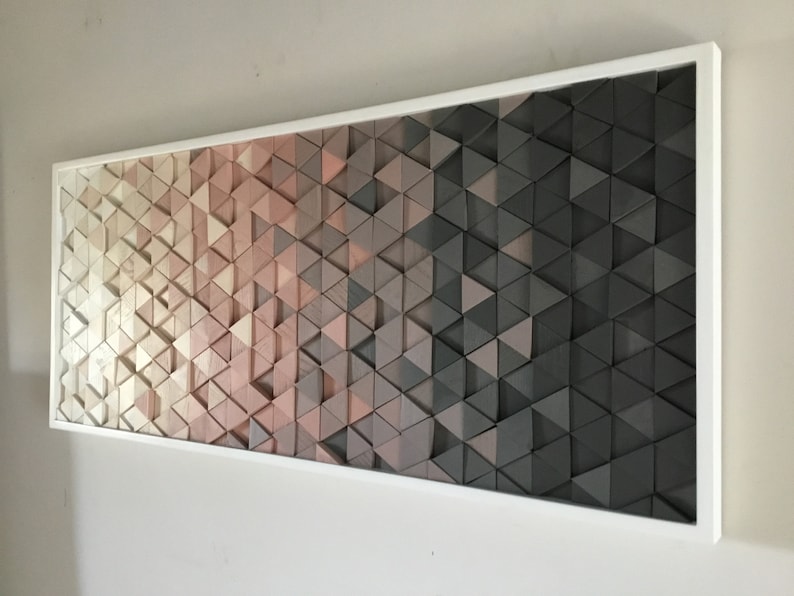 May include: A framed wall art piece made of wood triangles in shades of white, pink, and gray. The triangles are arranged in a gradient pattern, creating a modern and abstract design.
