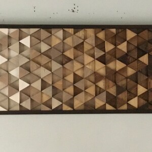 May include: A rectangular wooden wall art piece with a geometric pattern of triangles in shades of brown, beige, and white. The triangles are arranged in a gradient pattern, with the lightest shades at the top and the darkest shades at the bottom.